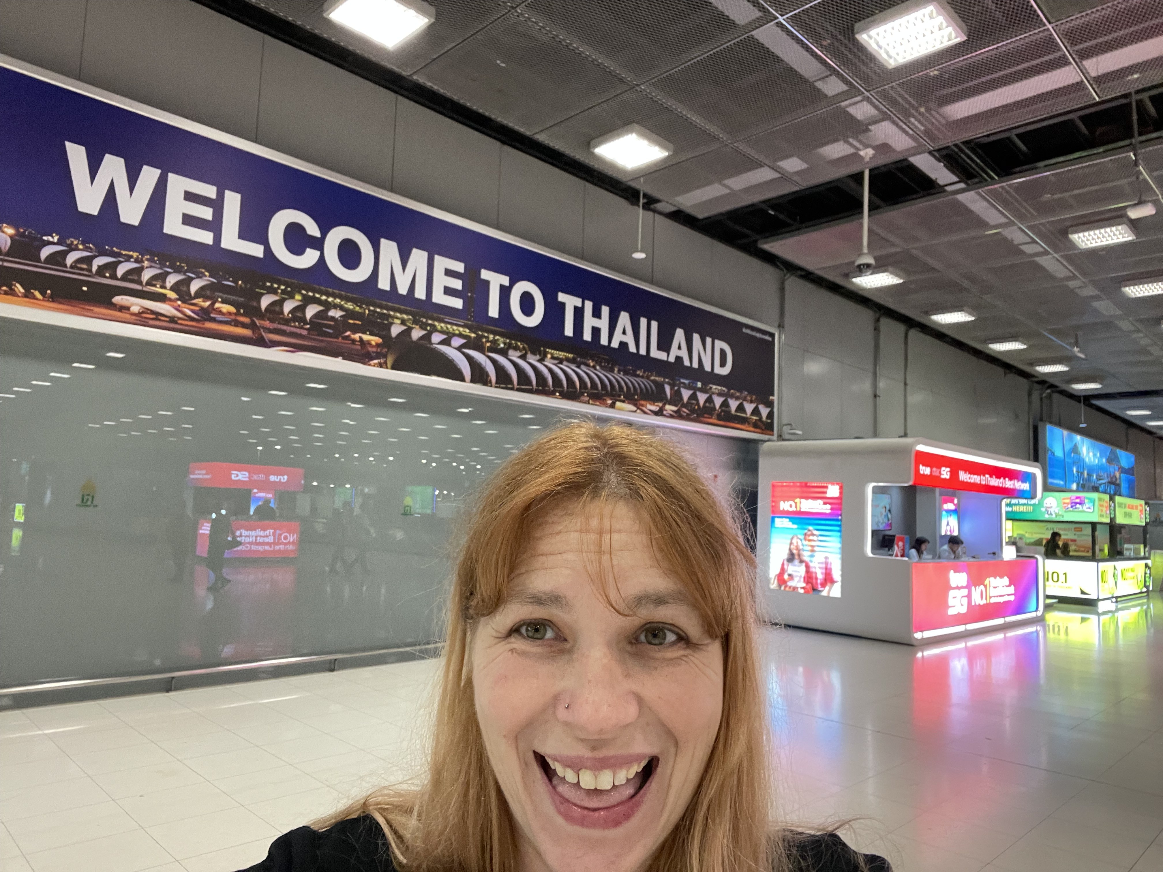 Mari smiling in the Bangkok airport under a Welcome to Thailand sign, the day she arrived